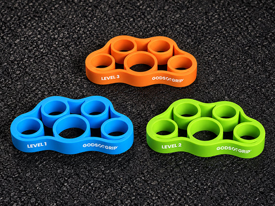 Three God of Grip finger grip trainers in orange, blue, and green, arranged on a dark textured asphalt surface. The orange one is labeled Level 3, the blue one Level 1, and the green one Level 2.