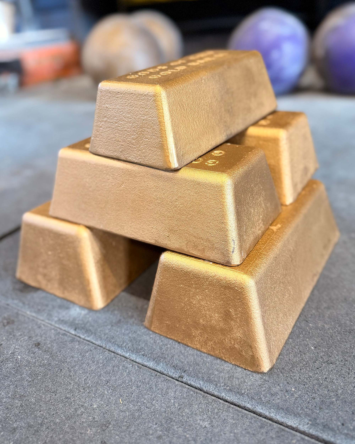 Five stacked gold-colored metal bars with a textured finish, photographed on a gray surface with a blurred background.