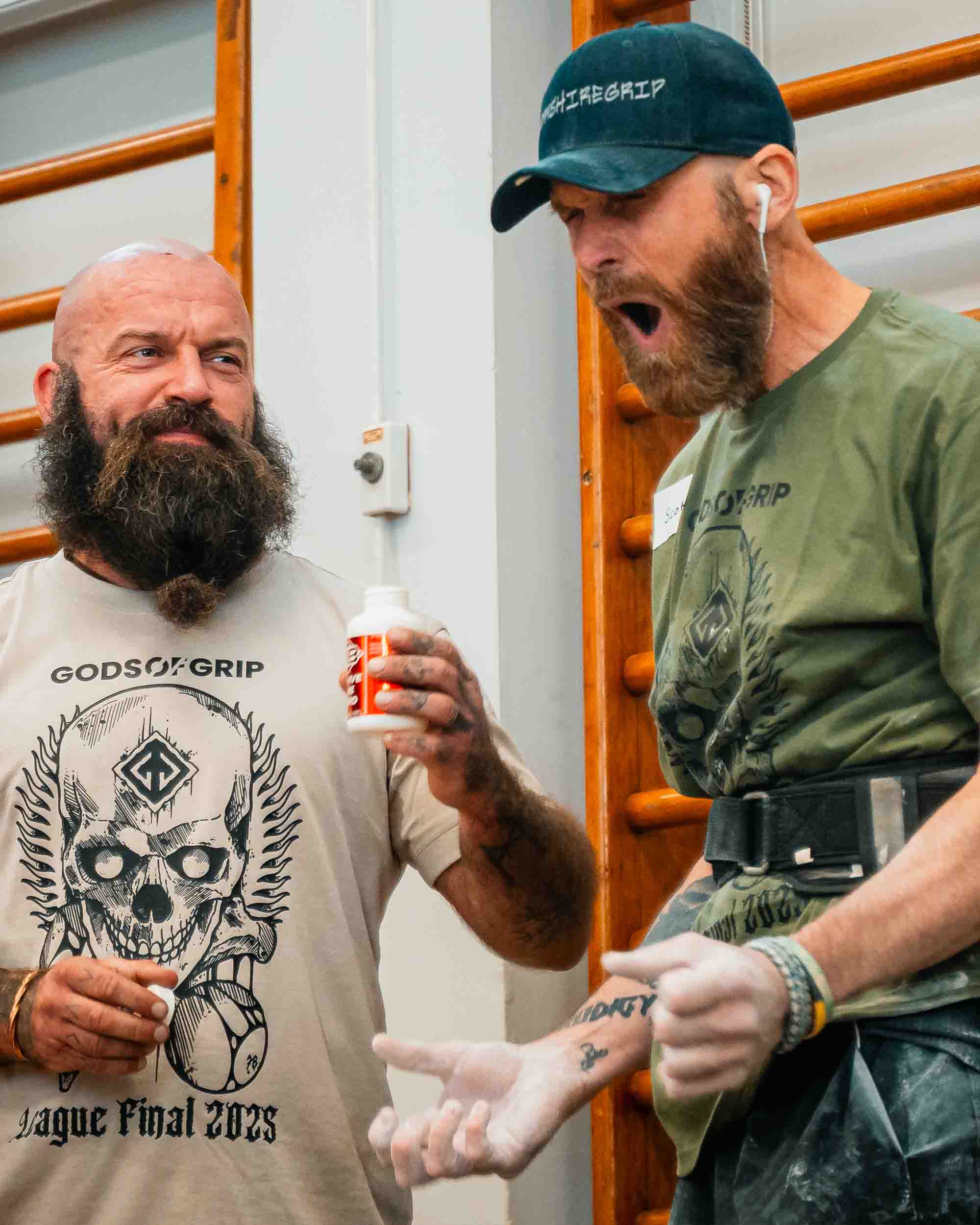 Two men, one holding a bottle, standing indoors with wooden railings in the background.