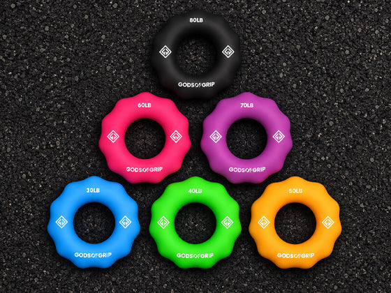 Six God's Grip hand grip trainers in different colors (black, pink, purple, blue, green, orange) arranged in a pyramid on a dark asphalt background. Each grip trainer is labeled with its weight capacity (30LB, 40LB, 40LB, 50LB, 60LB, 70LB) and the God's Grip brand logo.